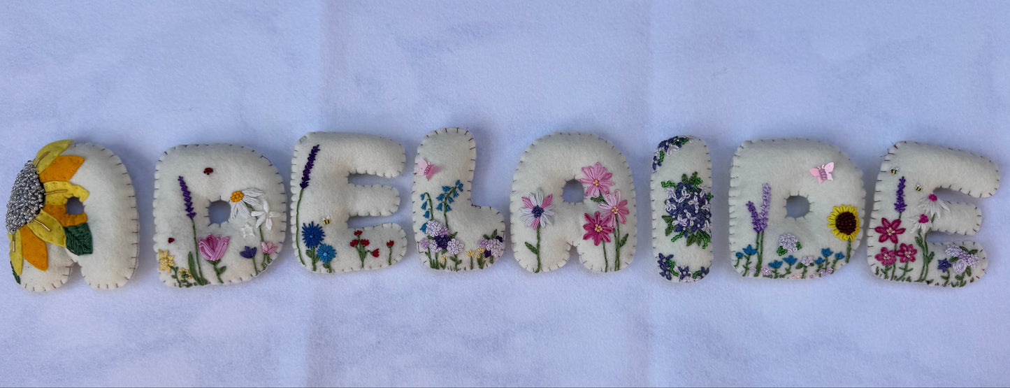 Eight embroidered letters spelling 'ADELAIDE' arranged vertically on a light blue textured fabric background. Each letter is made from off-white fabric with colorful floral and botanical embroidery.