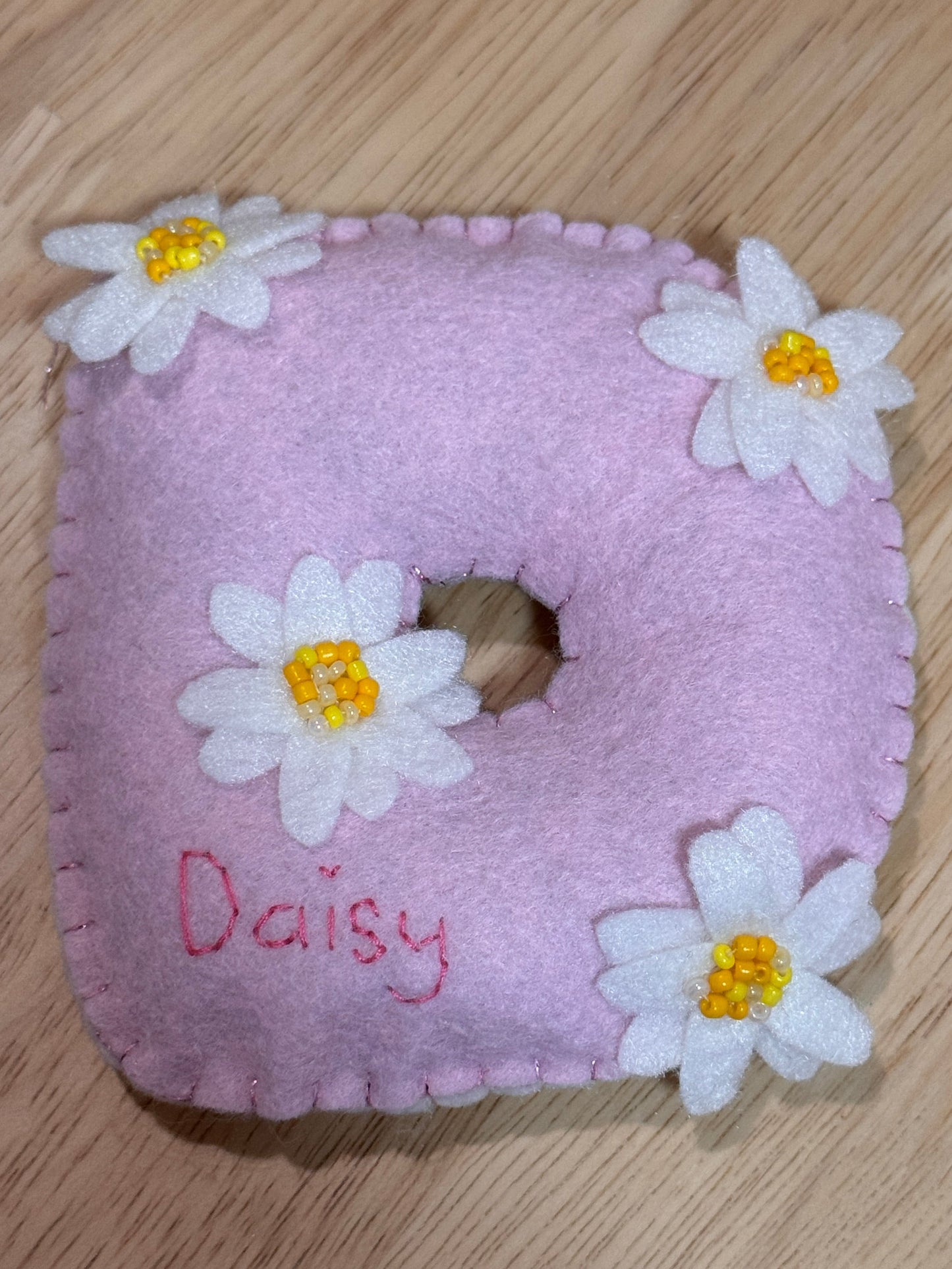 A handmade pink felt  Dshaped item with embroidered white daisy flowers with yellow centers and the name 'Daisy' stitched in pink, shown on a light wood surface.