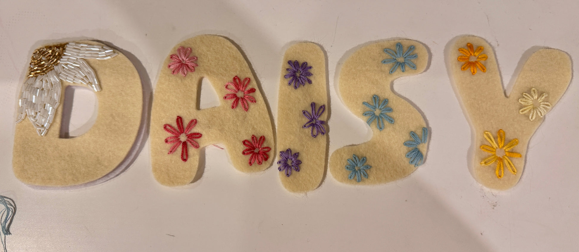 Felt letters spelling 'DAISY' arranged on a white surface, with embroidered flowers in pink, purple, blue, yellow, and white, plus a small decorative embellishment on the letter D.