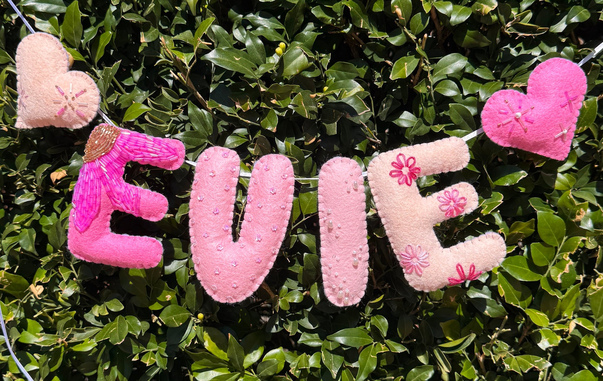 Personalized felt letter banner spelling 'EVIE' with pink and beige letters, hanging outdoors against a green hedge background.