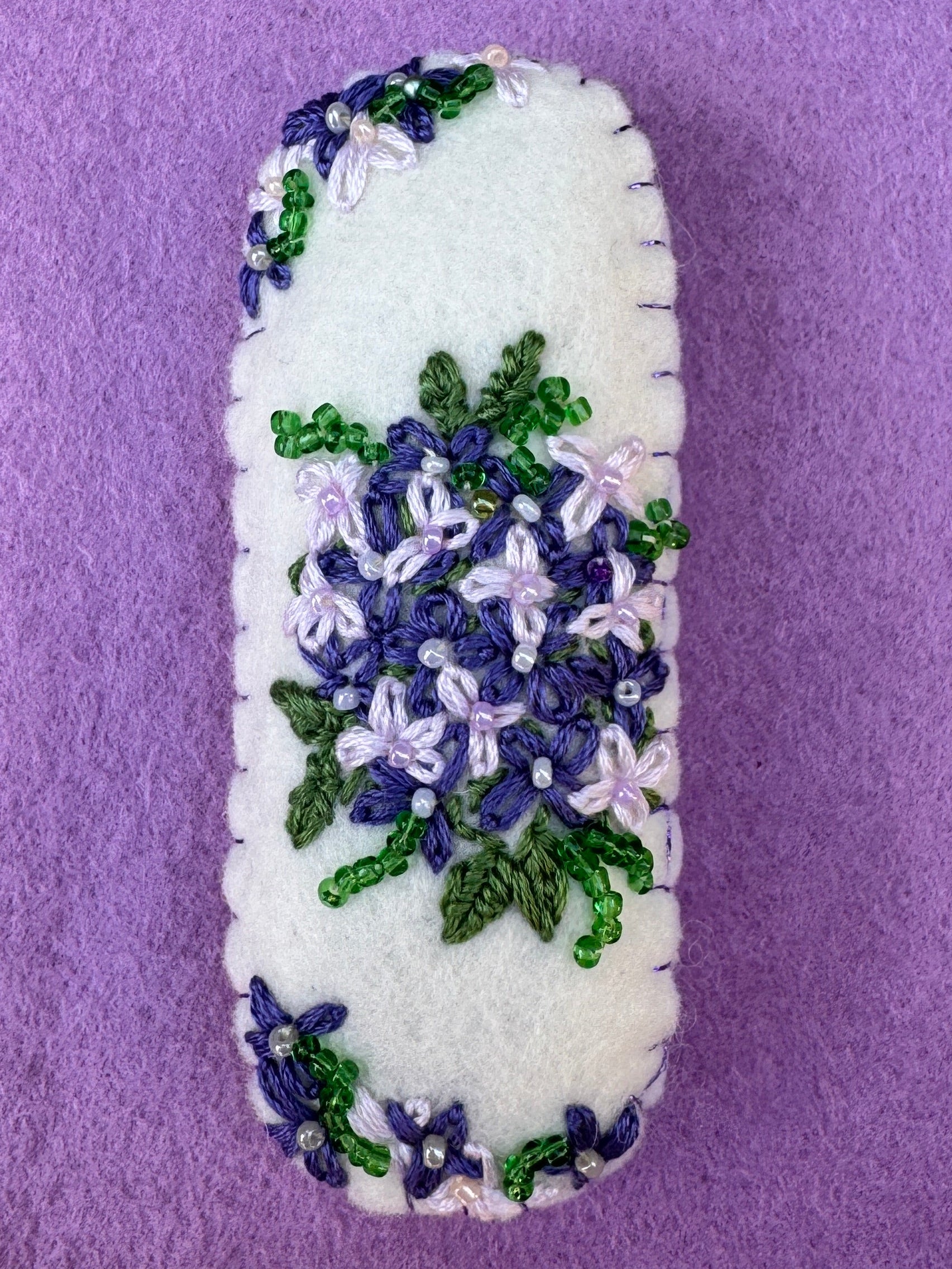 A small white embroidered letter I, featuring a floral pattern in purple, blue, and green, displayed on a textured purple background.