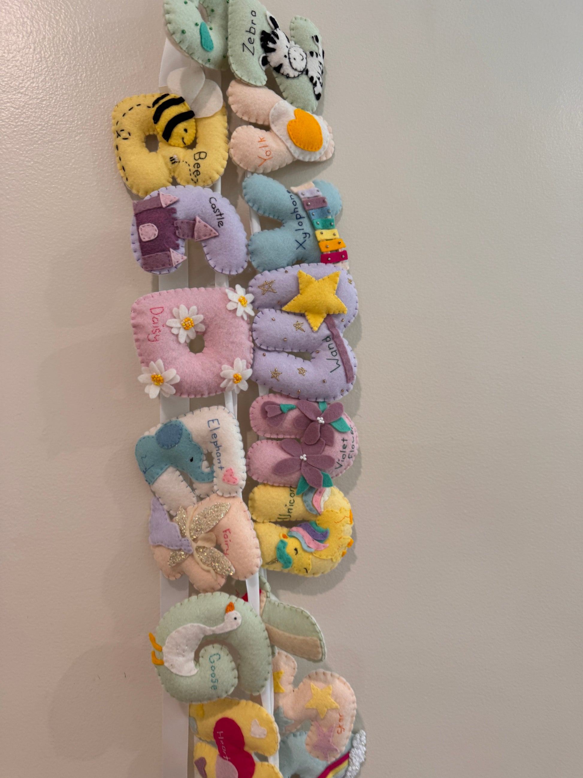 A vertical arrangement of colorful, soft fabric letters, featuring various shapes like a bee, moon, flower, and star in pastel and bright colors against a plain light background.