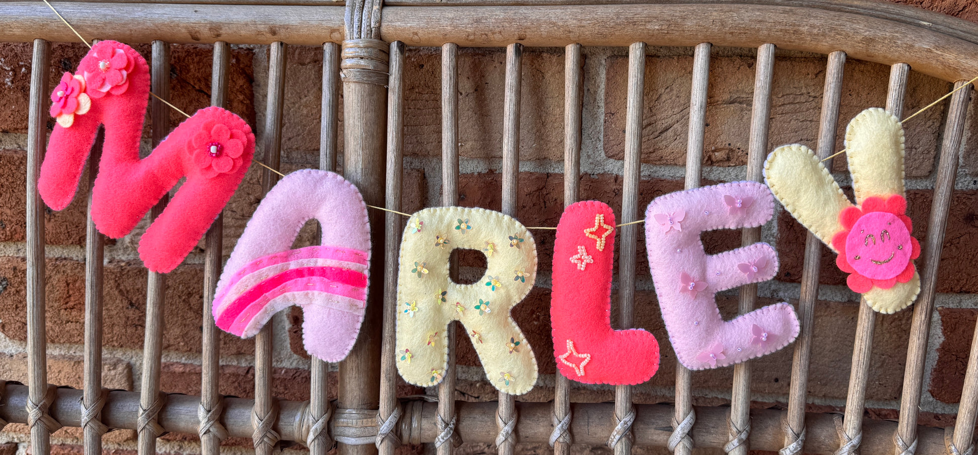 Personalized felt letter banner spelling 'MARLEY' hanging on a rope in front of a rustic wooden slatted background. The letters are made from pink, lavender, and cream fabrics with embroidered details and small flower accents.