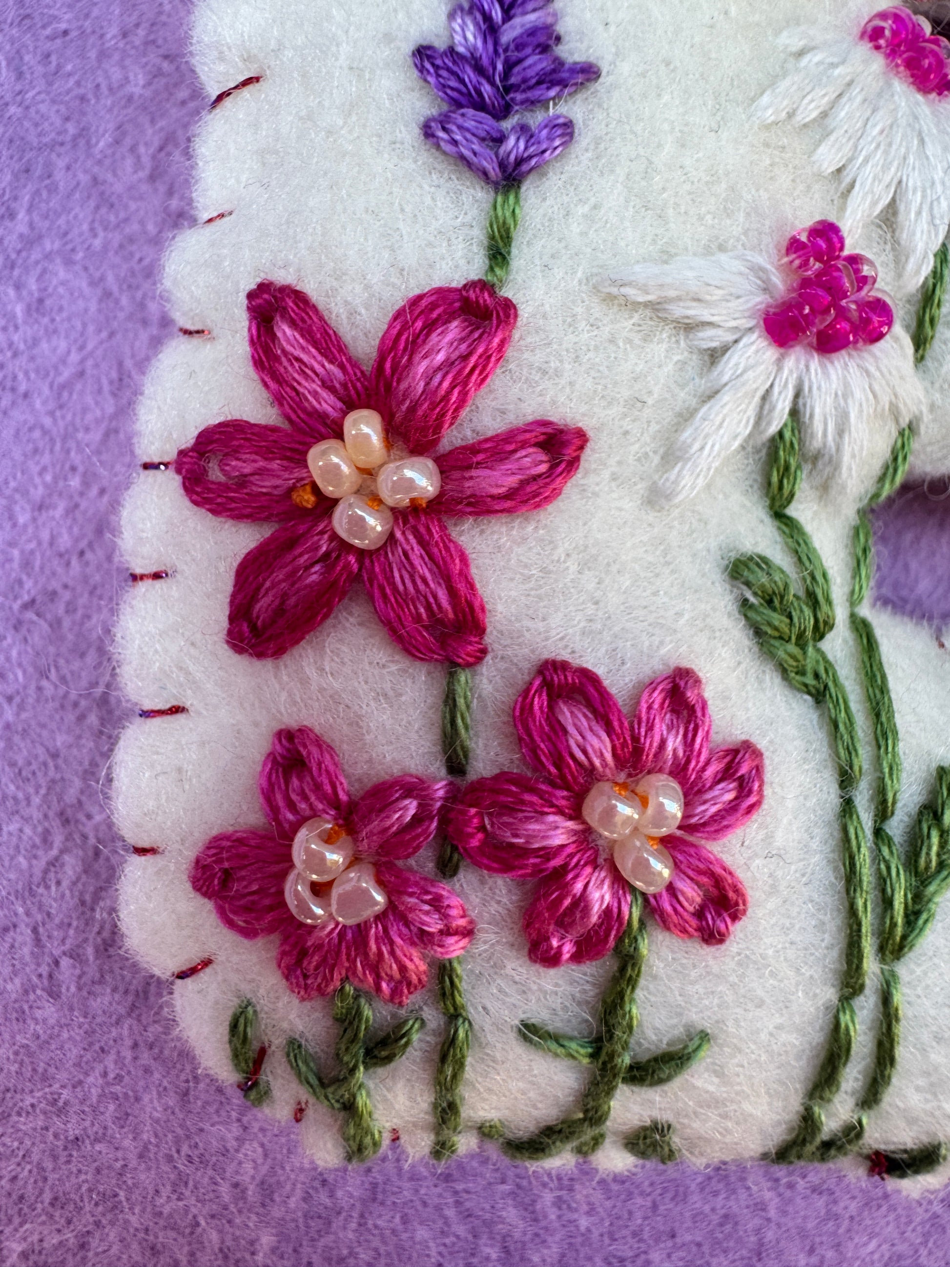 Close-up of a white embroidered felt letter, featuring pink and purple flowers with green stems and pearl-like centers, set against a purple background.