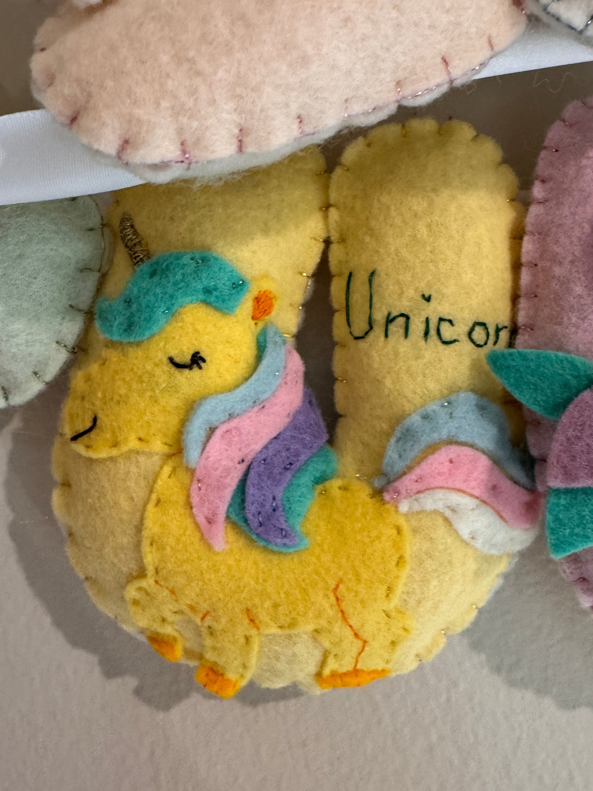 A close-up of handmade felt letter including a yellow unicorn-with a pastel rainbow mane and the word 'Unicorn' stitched on it, alongside other pastel felt shoes.