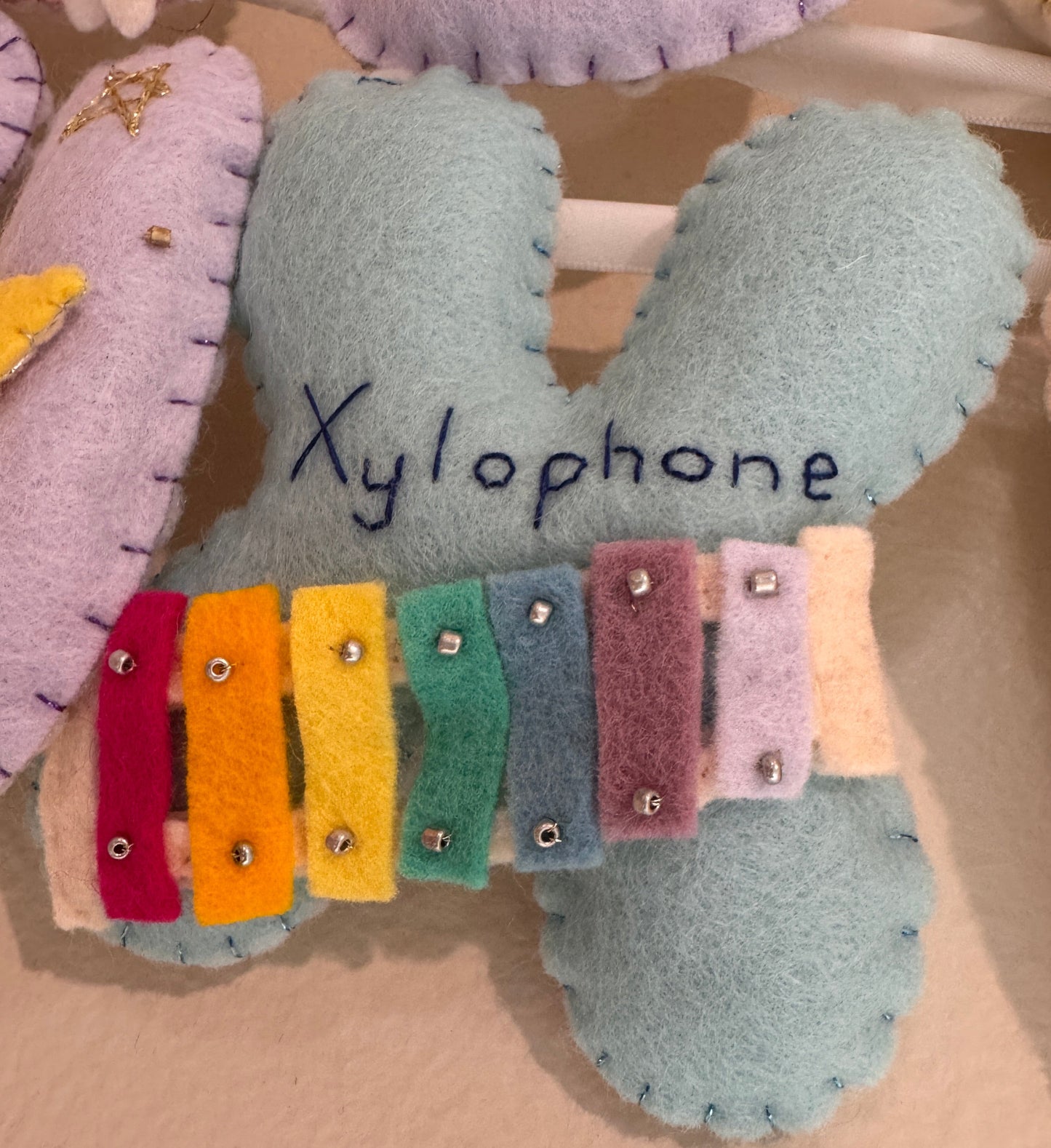 A handmade felt baby xylophone letter, featuring pastel blue-green and colorful felt keys in yellow, orange, red, pink, green, blue, purple, and cream, with the word 'Xylophone' embroidered on the side.