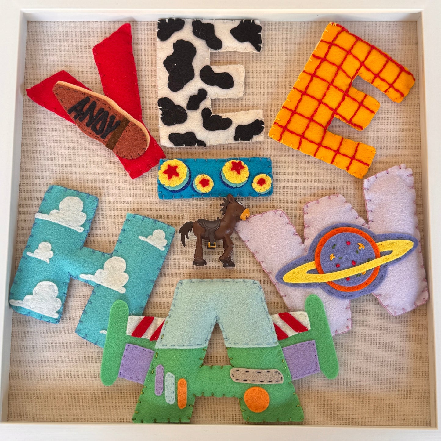 A framed felt shadow box featuring the word 'YEE-HAW' in colorful, Toy Story themed letters. The letters spell out 'YEE-HAW' using various pop culture and sci-fi motifs: a cow-print 'E', a grid-patterned 'H', a red 'L' shaped like a taco with 'ARMY' stitched inside, a blue 'L' with clouds, a green 'H' with spaceships, and a purple 'O' with a ringed planet.